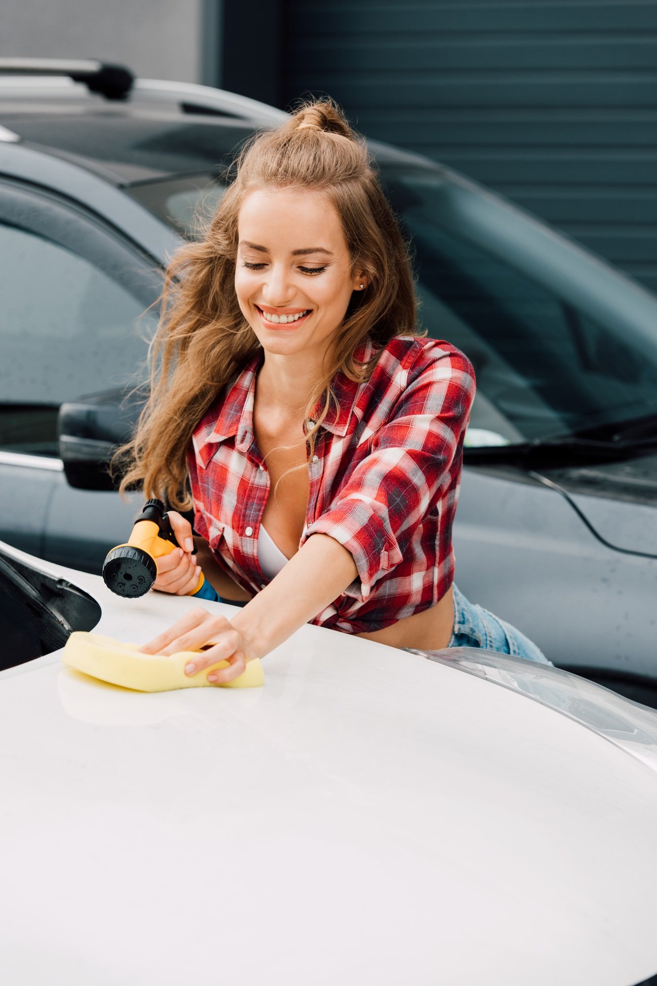 happy girl holding pressure washer and sponge while washing car