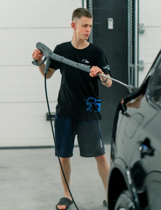 A male car wash employee washes a black luxury car with high-pressure washer in the car wash bay A male car wash employee washes a black luxury car with high-pressure washer in the car wash bay